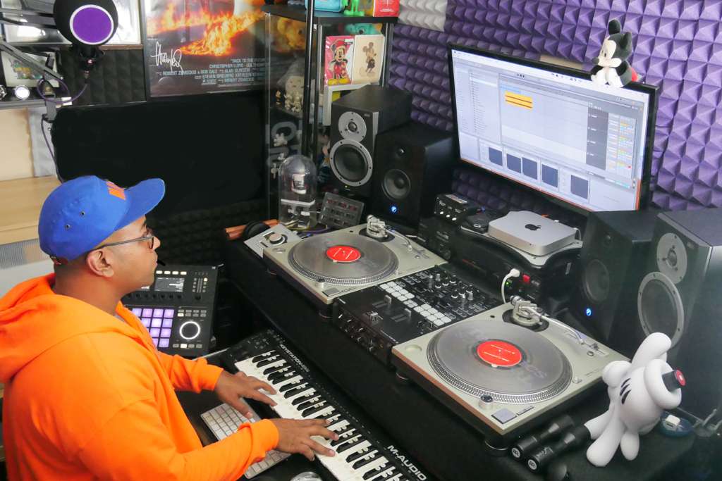 Hanif Esmail in his studio, playing a keyboard beside a pair of turntables and a mixer, facing a screen and studio monitors in an acoustically treated room.